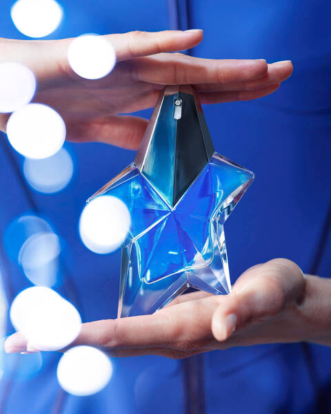 Two hands holding Mugler Angel eau de parfum shooting star bottle from top and bottom on blue background with blurry lights