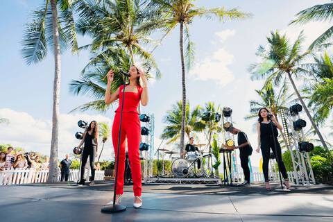 Singer Dua Lipa on stage in red Mugler outfit with white stillettos on stage with band surrounded by palm trees