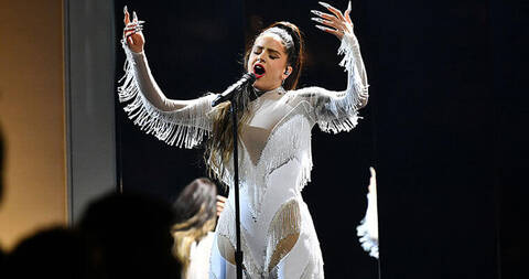 Singer Rosalia in white fringe Mugler outfit with high pony tail singing into standup microphone