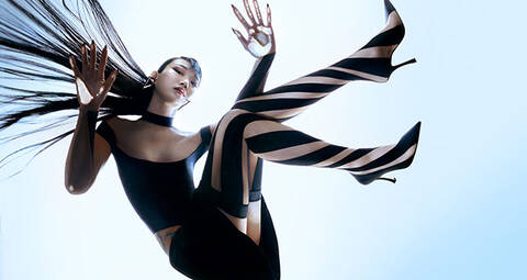 Model photographed from below glass floor wearing striped tights, cropped black top and black skirt, with long fanned out hair
