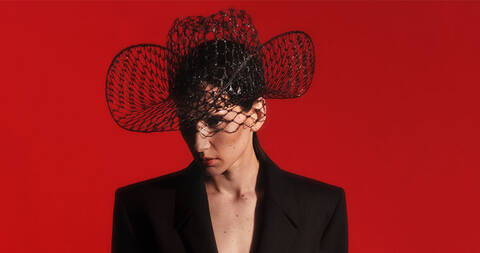 Bust shot of model wearing mesh cowboy hat and black blazer on red background