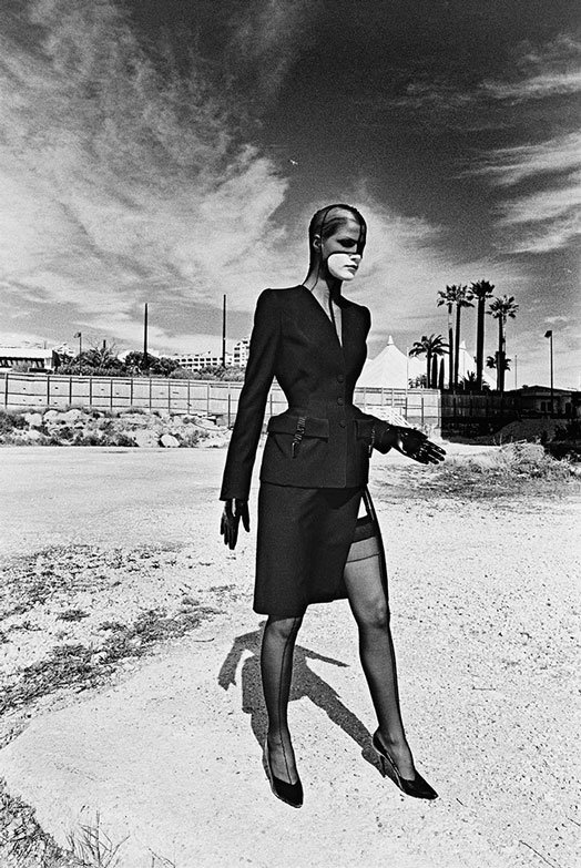 A black and white photo of a woman wearing a skirt suit, leather gloves, stockings, and a stocking headpiece